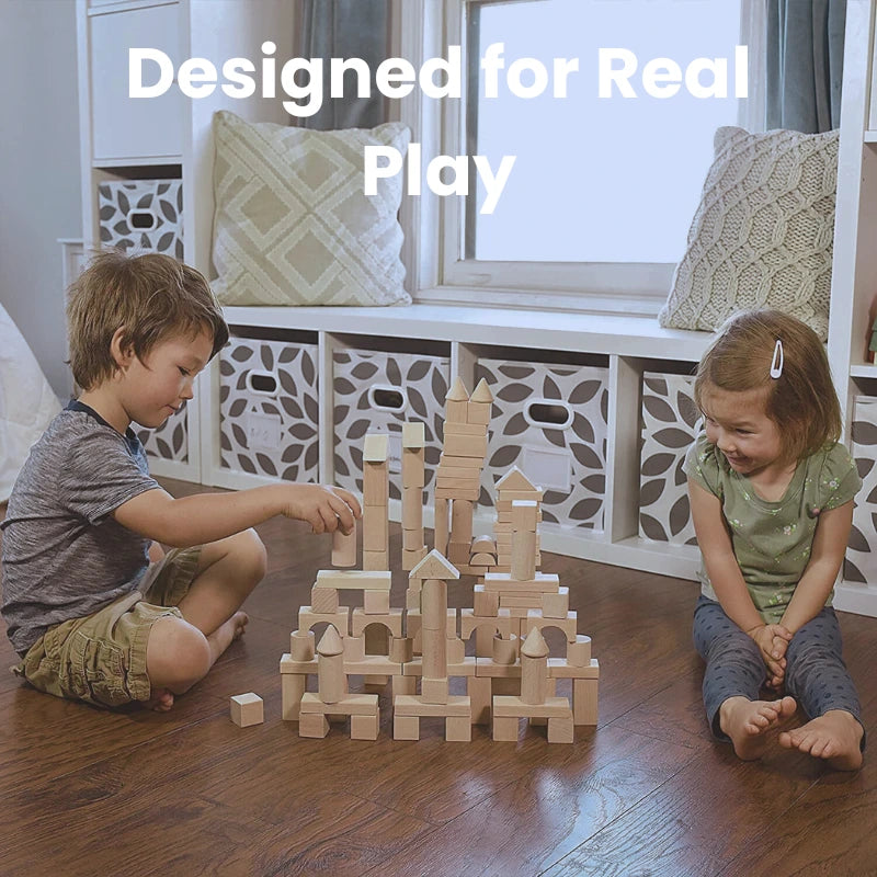 Two children sitting on a wooden floor, building a tall castle-like structure with natural wood building blocks. The setting includes a cozy room with storage bins and pillows, showcasing collaborative play for toddlers.