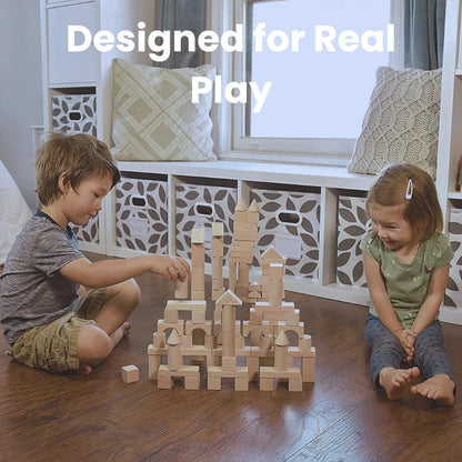 Two children sitting on a wooden floor, building a tall castle-like structure with natural wood building blocks. The setting includes a cozy room with storage bins and pillows, showcasing collaborative play for toddlers.